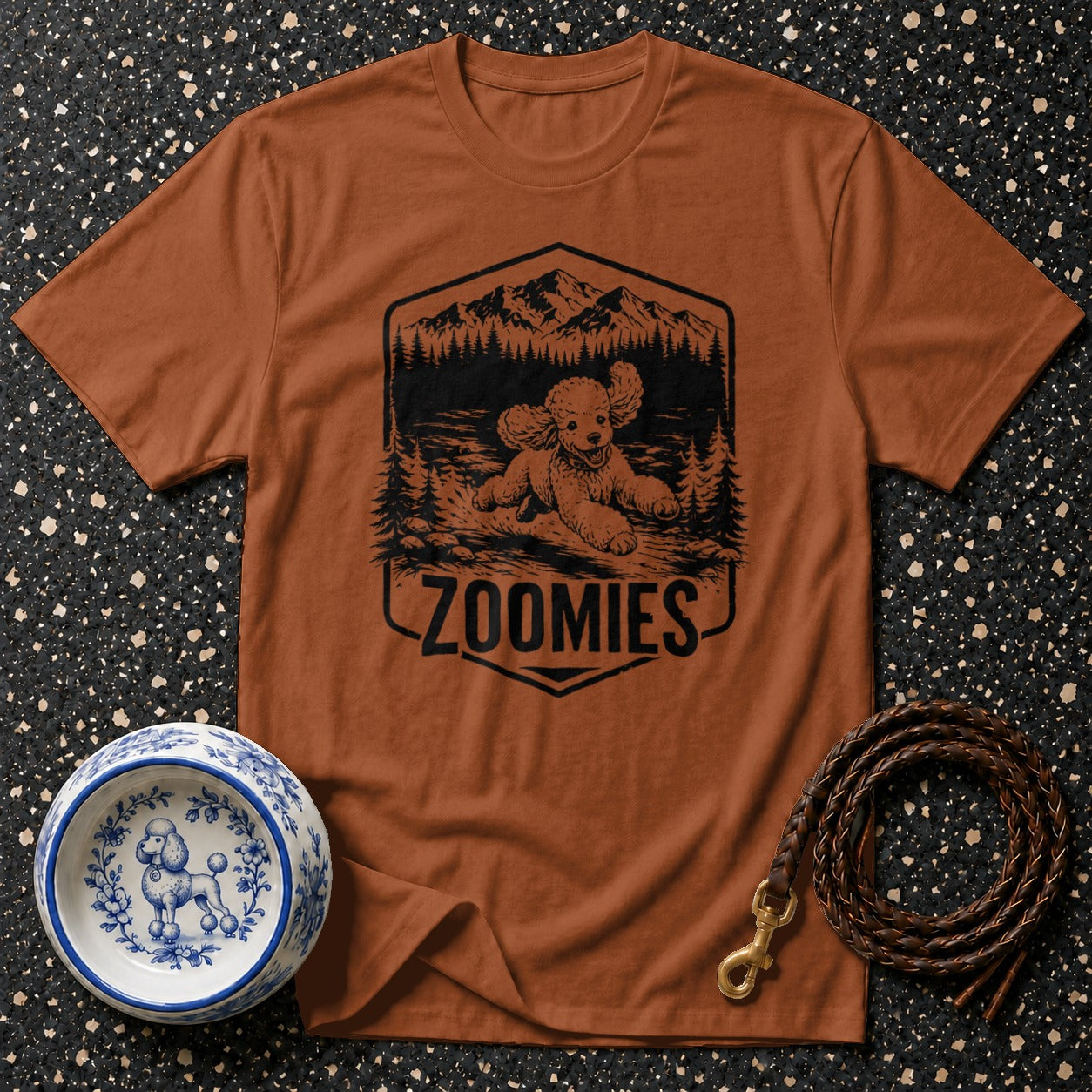 Brown t-shirt with a graphic of a poodle running through a forest with mountains and the word "ZOOMIES".