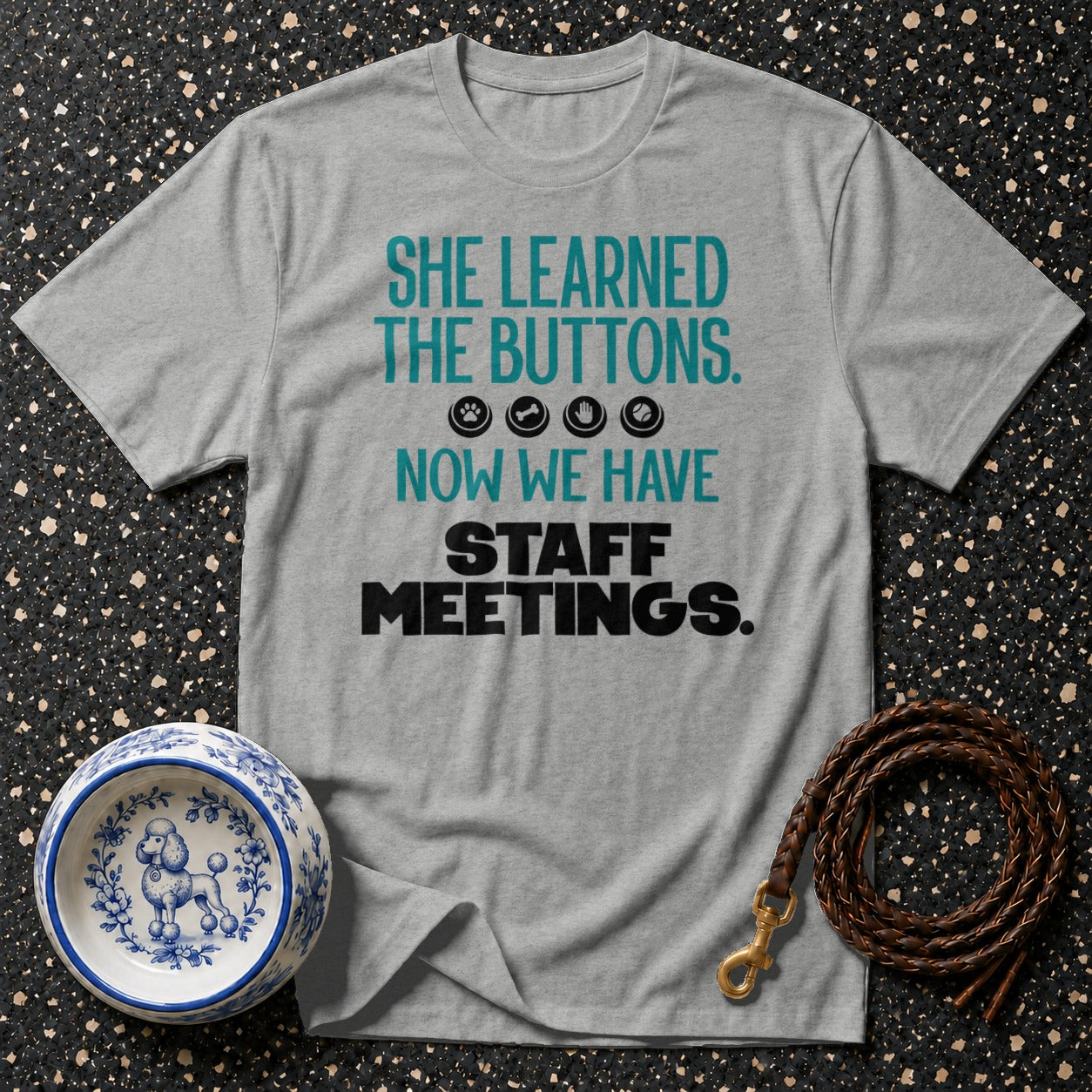Grey t-shirt with text: "SHE LEARNED THE BUTTONS. 🐾🦴🎾 NOW WE HAVE STAFF MEETINGS."