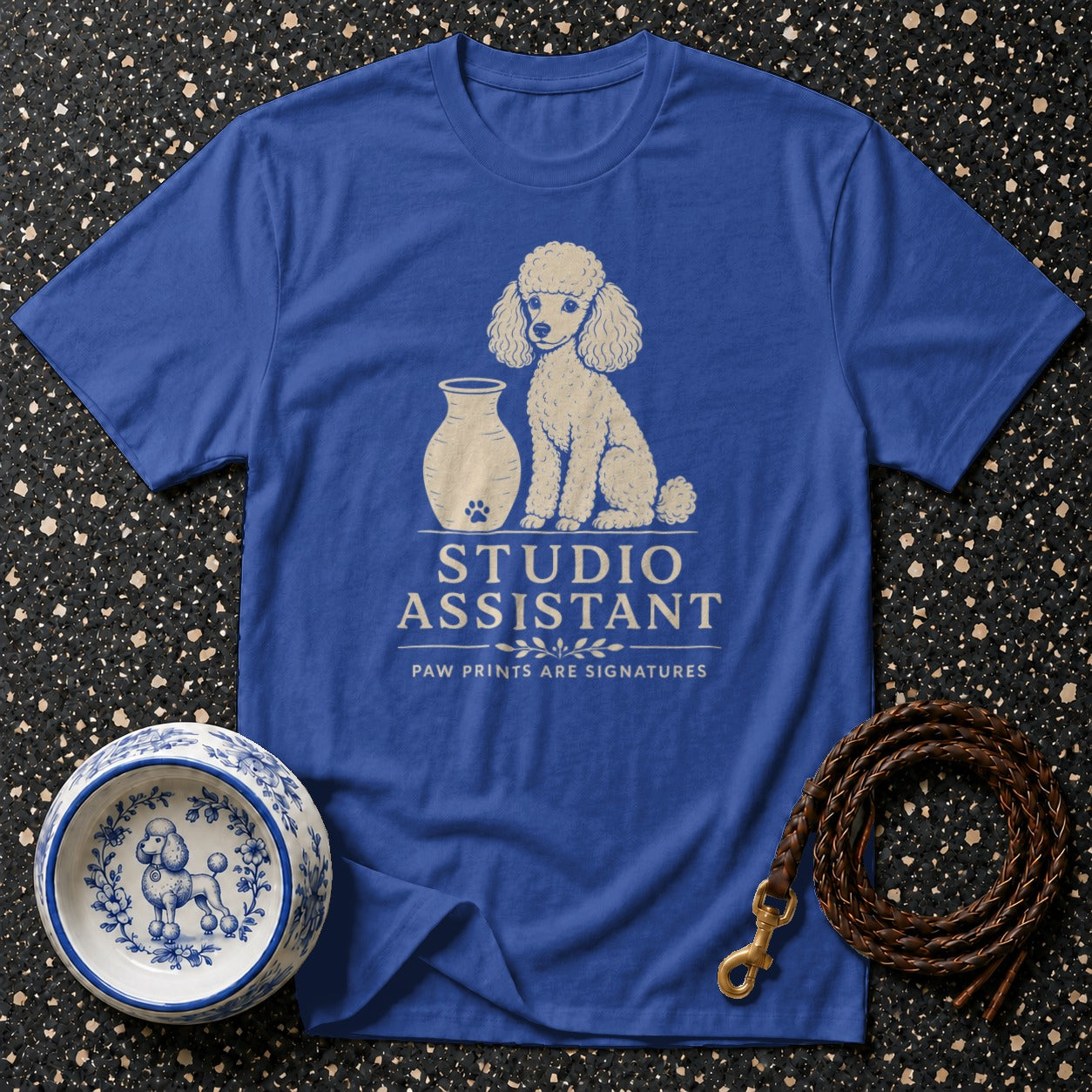 Blue t-shirt with a cream poodle and vase graphic. Text reads "STUDIO ASSISTANT PAW PRINTS ARE SIGNATURES".